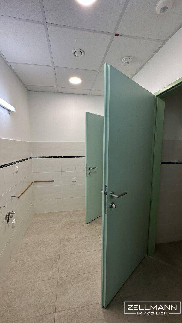Öffentlicher Waschraum mit mehreren Toilettenkabinen, hellgrünen Türen und weiß gefliesten Wänden mit schwarzem Akzentstreifen.