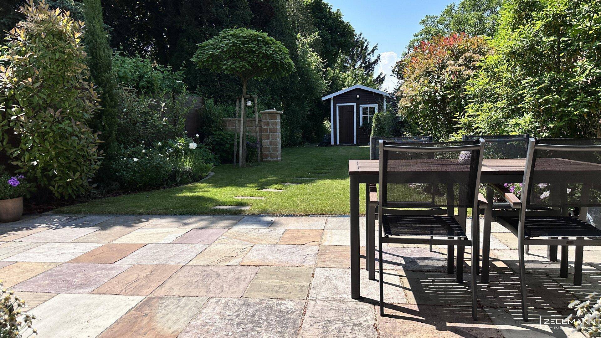 Sonniger Terrassenbereich mit Esstisch und Stühlen, angrenzend an einen gepflegten Garten.