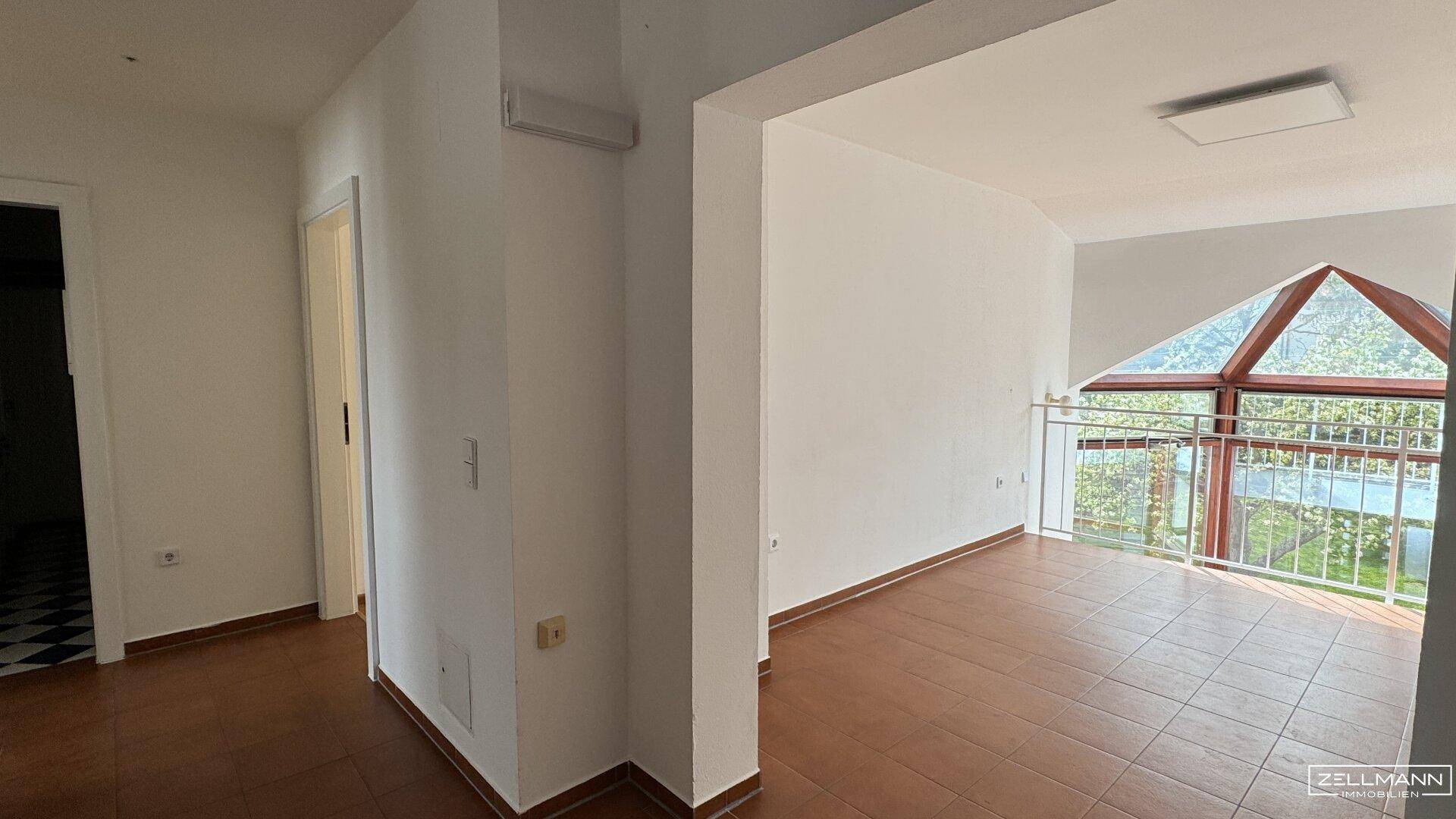 Heller Flur mit Fliesenboden, der zu einem Bereich mit großen, schrägen Fenstern und Balkon führt.
