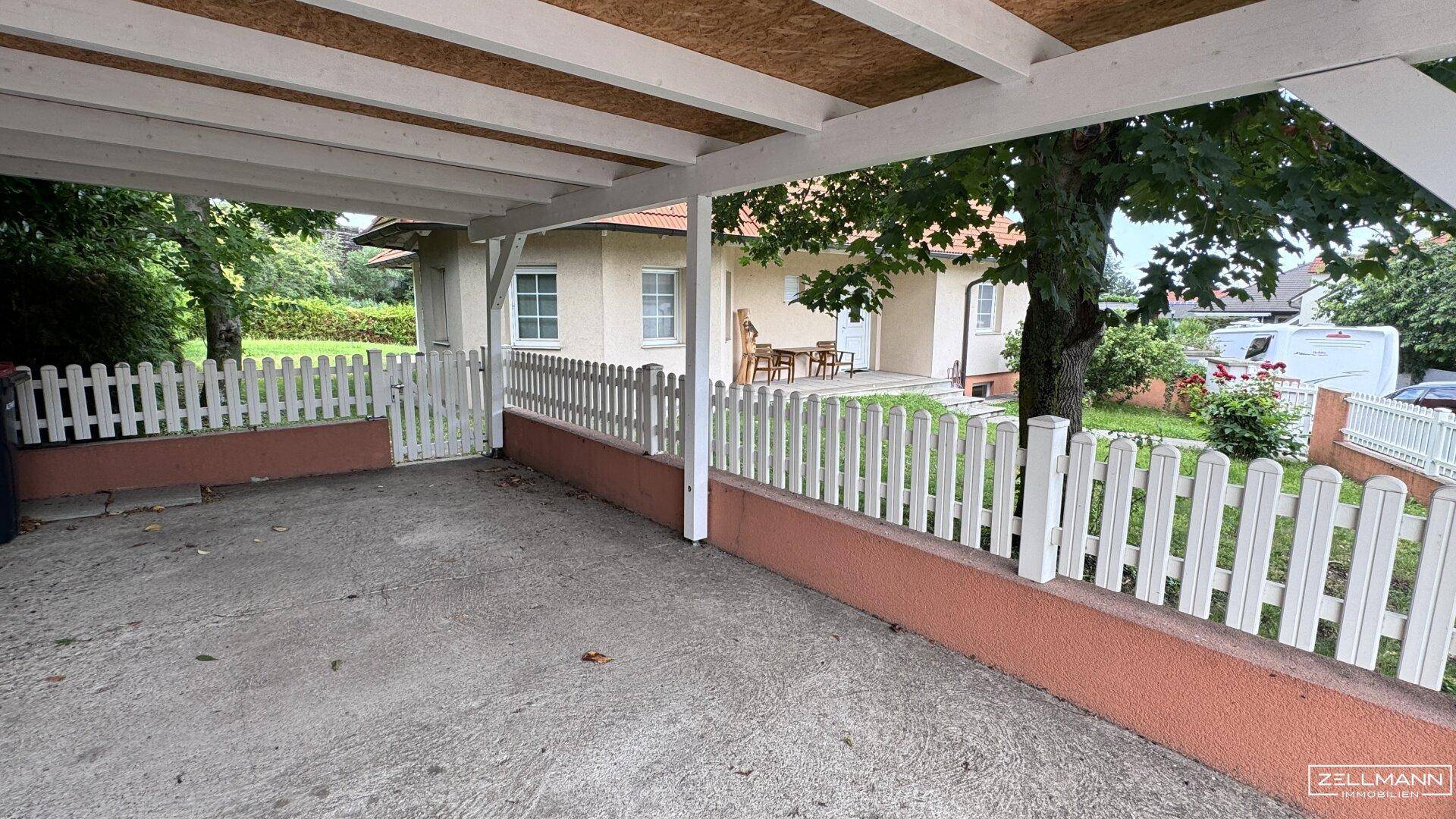 Überdachter Carport und Vorgarten mit weißem Zaun, bietet Schutz für Fahrzeuge.