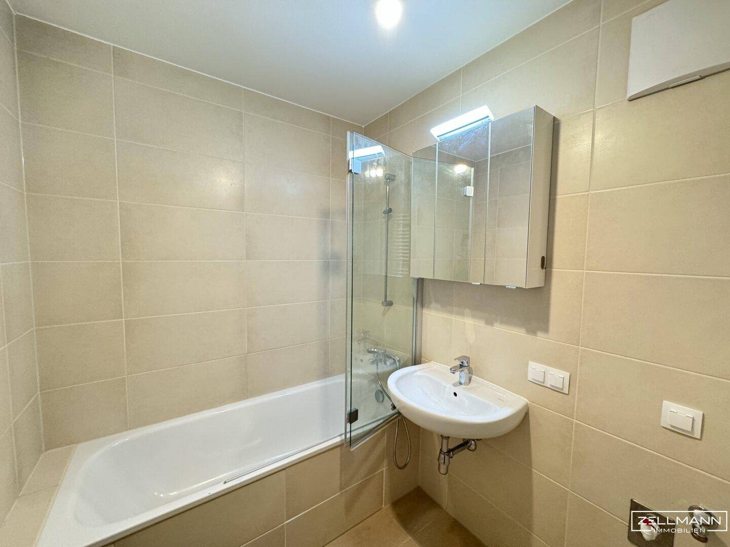 Helles Badezimmer mit beigefarbenen Fliesen, Badewanne mit Duschwand und Waschbecken.