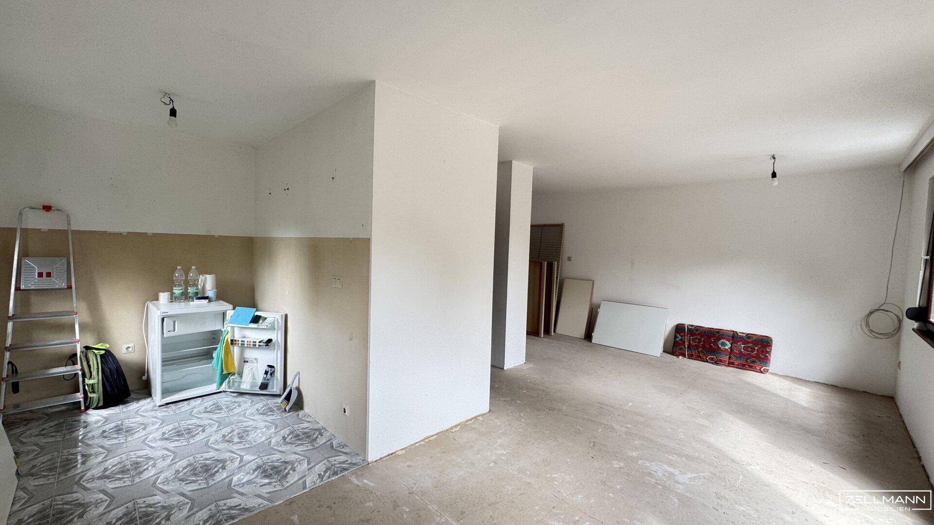 Unfertiger Wohnbereich mit kahlen Wänden, provisorischem Kühlschrank und Fliesenboden im Vordergrund.