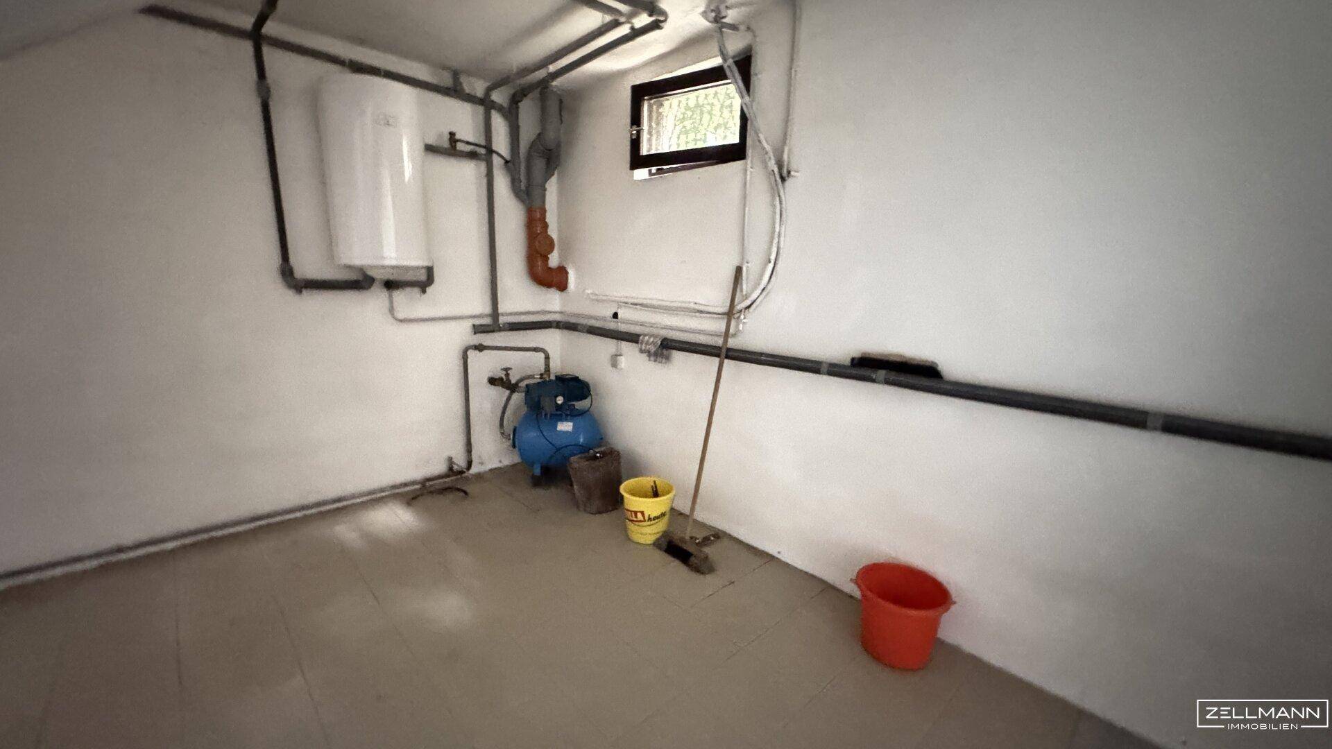 Technikraum im Keller mit Warmwasserboiler, sichtbaren Rohren und kleinem Fenster.