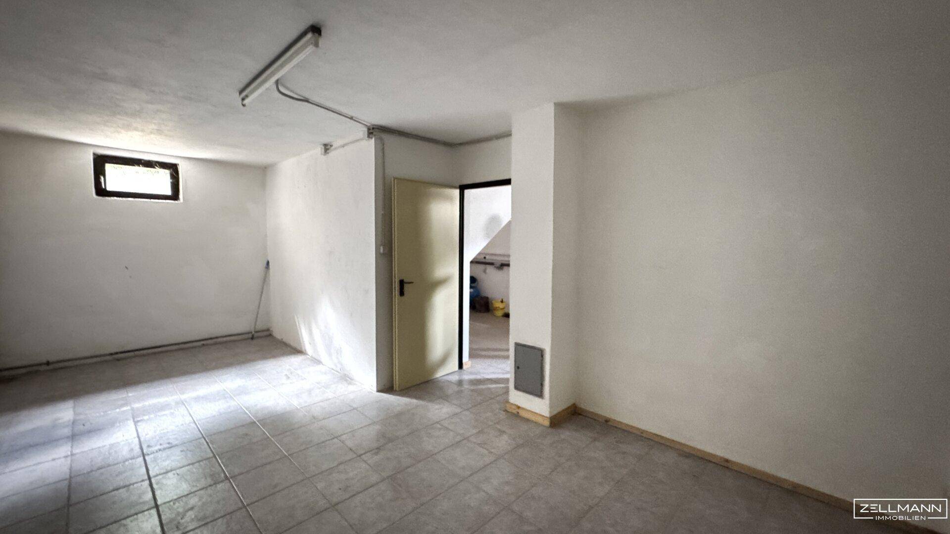 Heller Kellerraum mit Fliesenboden, kleinem Fenster und einfacher Deckenbeleuchtung.