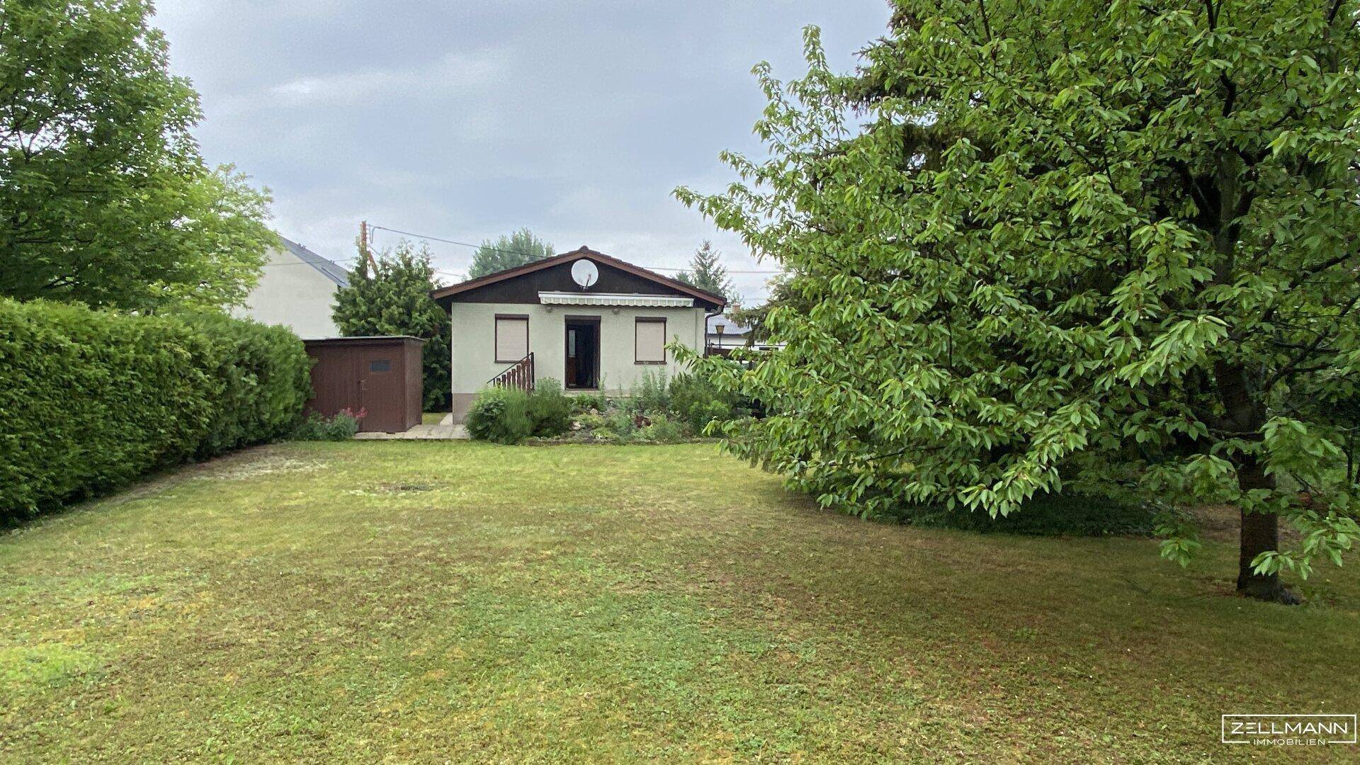 Gepflegter Garten mit Rasenfläche, Hecke und einem kleinen Einfamilienhaus im Hintergrund.