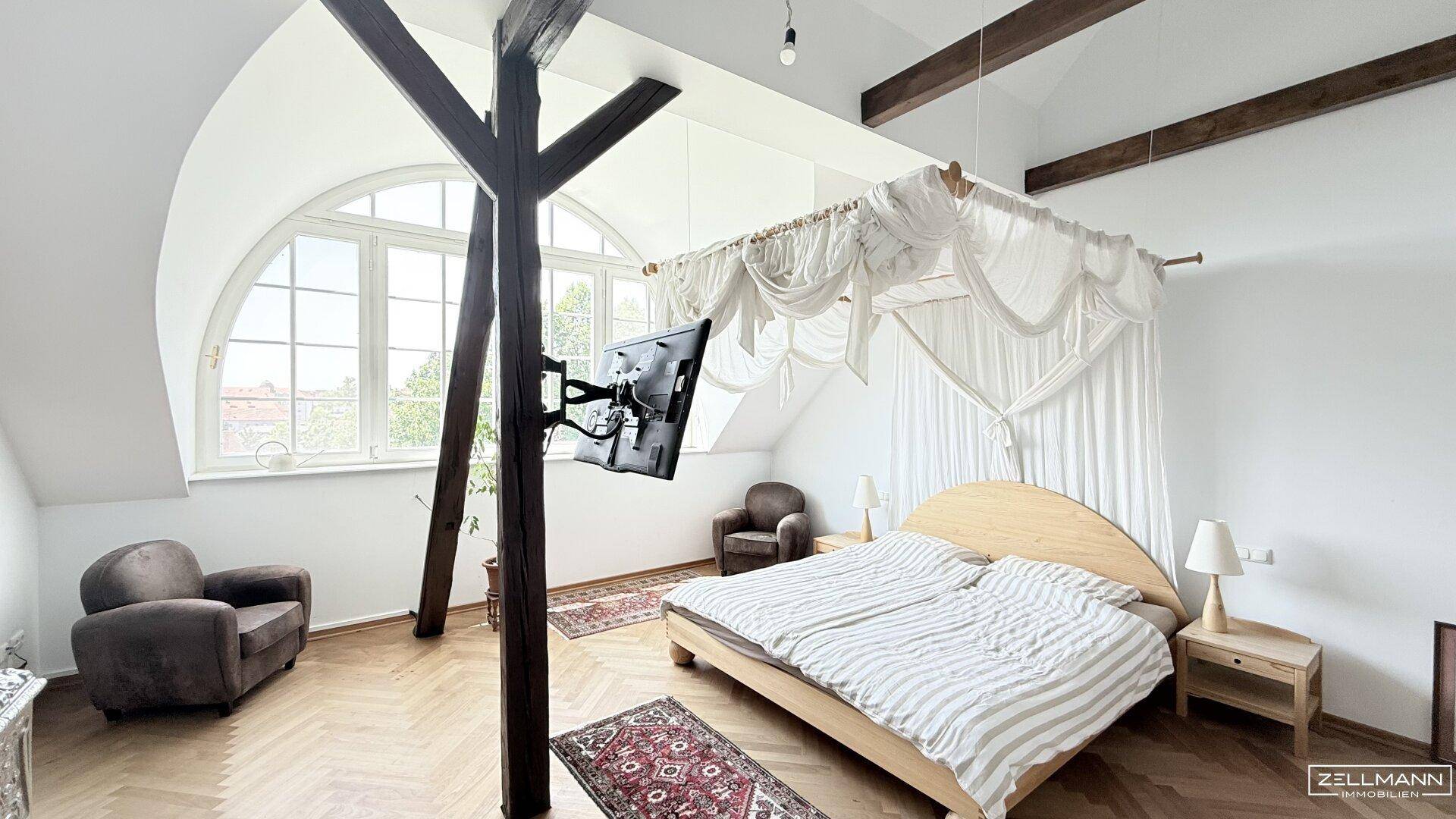 Geräumiges Schlafzimmer im Dachgeschoss mit Holzbalken, hohen Decken und großen Fenstern.