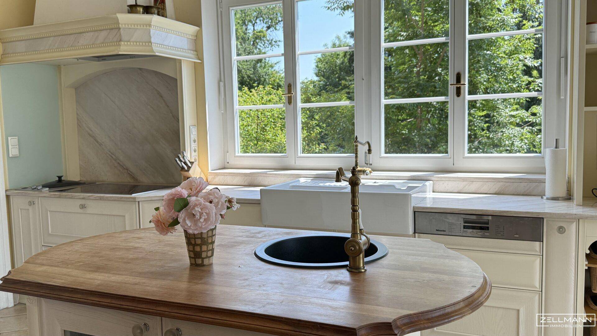 Helle Küche mit Holzinsel, Spülbecken und Blick ins Grüne durch große Fenster.