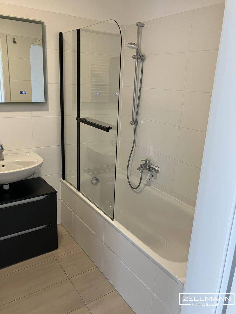 Modernes Badezimmer mit Badewanne, Duschwand, Waschbecken und hellen Fliesen.