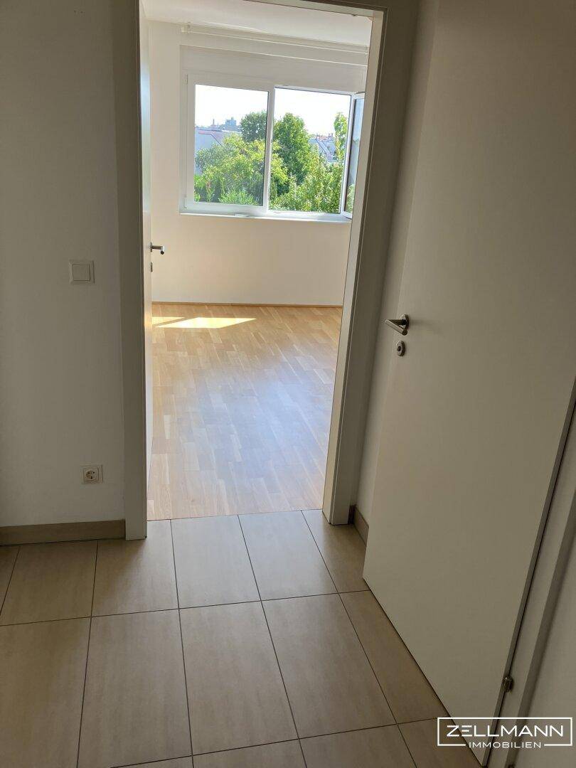 Flur mit Fliesenboden, der zu einem hellen Zimmer mit Holzboden und Fenster führt.