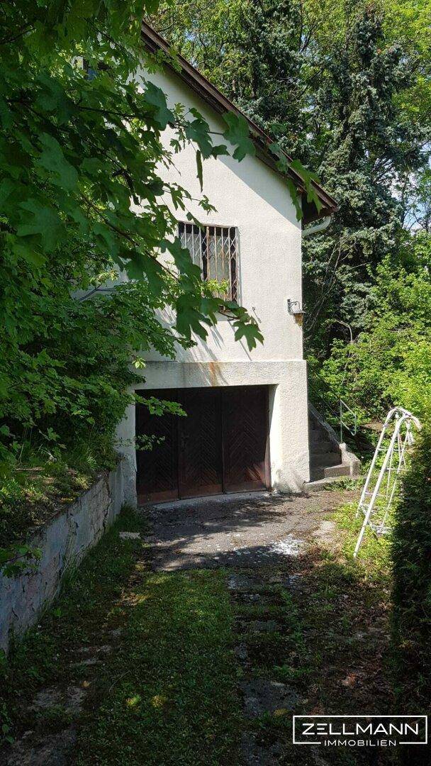 Die Fassade eines Hauses mit integrierter Garage und einem Fenster im oberen Stockwerk, umgeben von Bäumen.