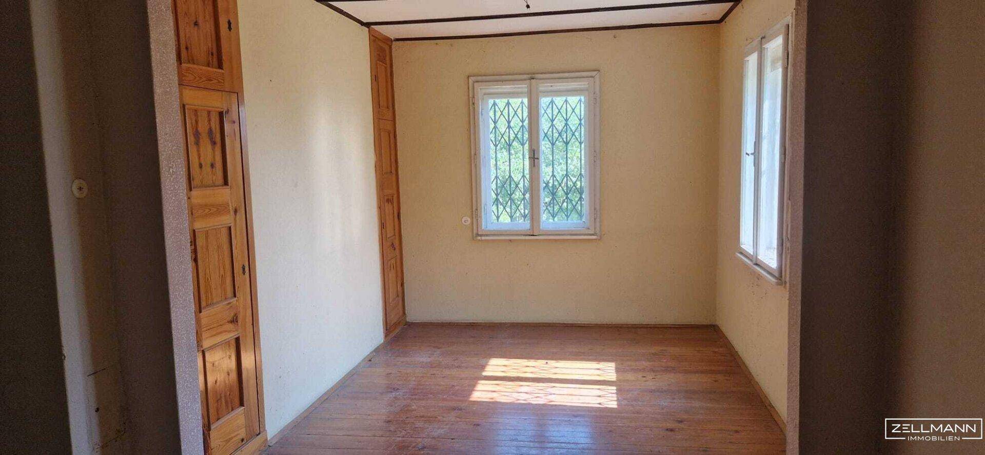 Leeres Zimmer mit Holzdielenboden, hellgelben Wänden und zwei Fenstern mit Sicherheitsgittern.