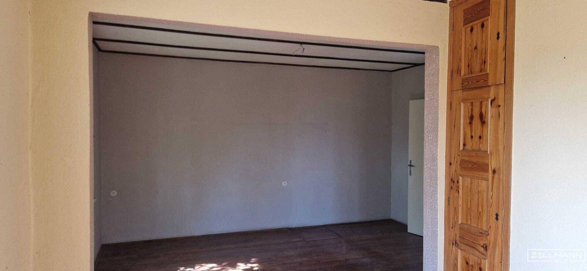 Leerer Raum mit Holzdielenboden und hellgrauen Wänden, gesehen durch einen breiten Durchgang.