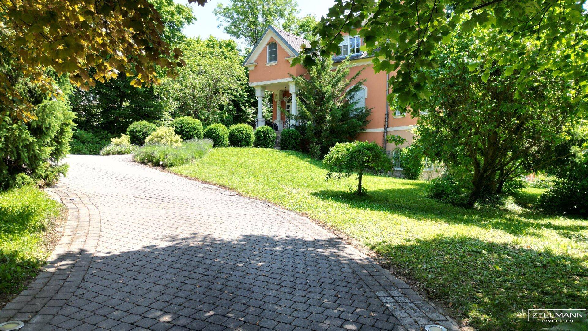 Gepflasterte Auffahrt, umgeben von einem gepflegten Vorgarten mit Bäumen und Sträuchern, führt zur Villa.