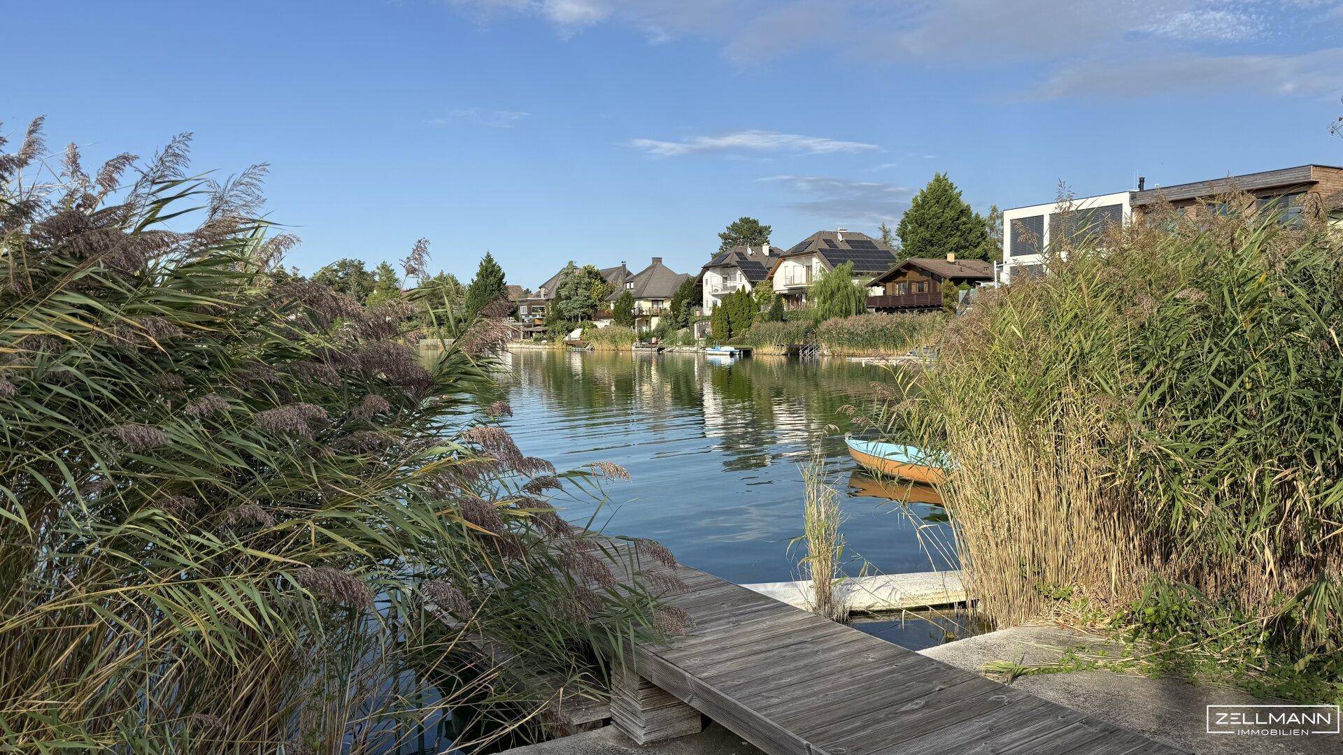 Malerischer Seeblick mit Häusern am Ufer, Schilf und einem kleinen Boot unter klarem Himmel.