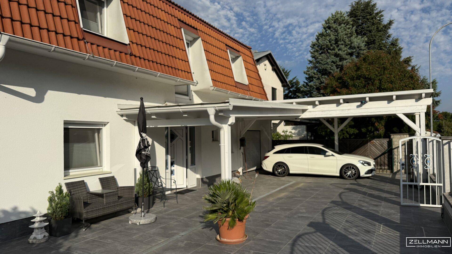 Vorderansicht des Hauses mit gepflasterter Auffahrt, Carport und einem kleinen Sitzbereich im Freien.