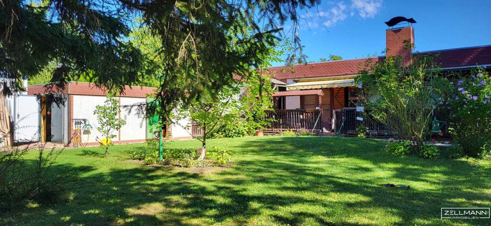 Gepflegter Garten mit Rasenfläche, Bäumen und einer Terrasse am Haus unter blauem Himmel.