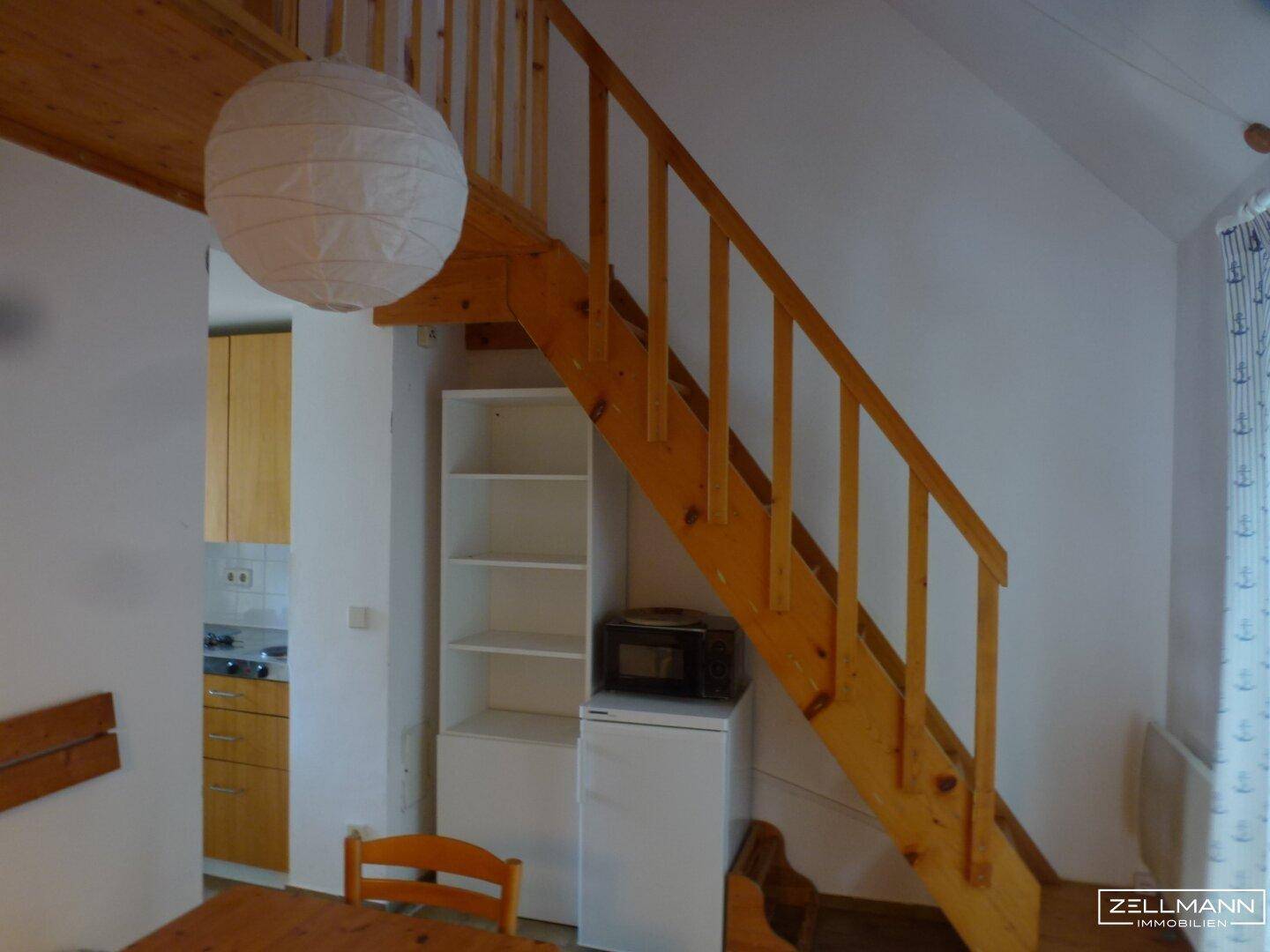 Holztreppe führt zur Galerie, darunter eine kleine Küchenzeile mit Mikrowelle und Kühlschrank.