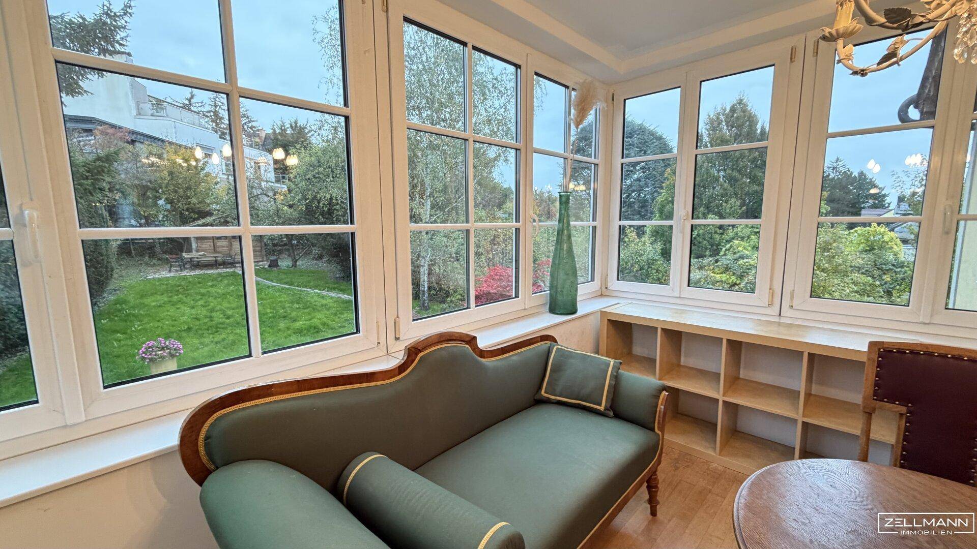 Heller Wintergarten mit vielen Fenstern, Blick in den Garten und einer eleganten Chaiselongue.