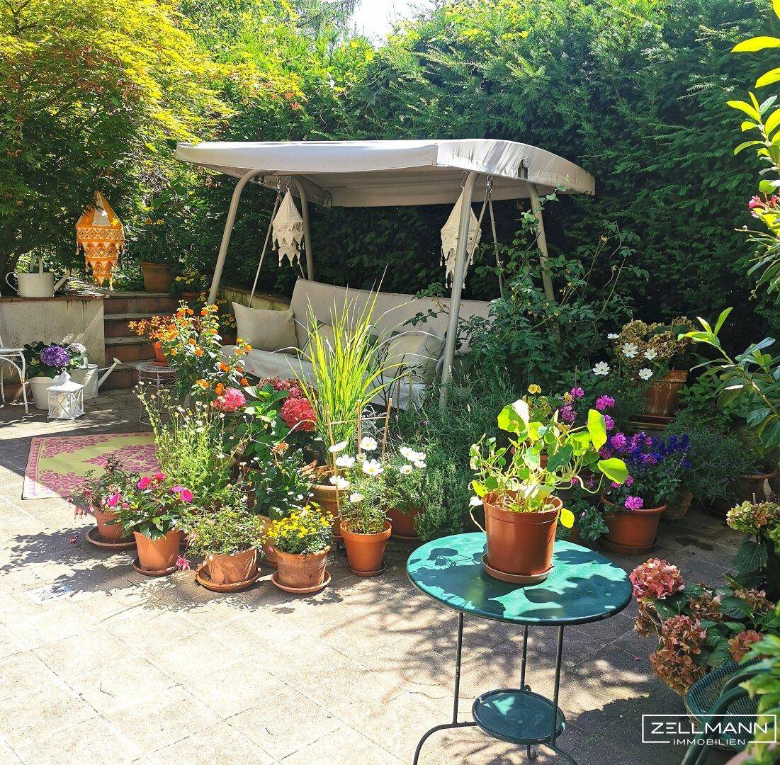 Sonnige Gartenterrasse mit Schaukelbank, vielen Blumen und üppiger grüner Bepflanzung.