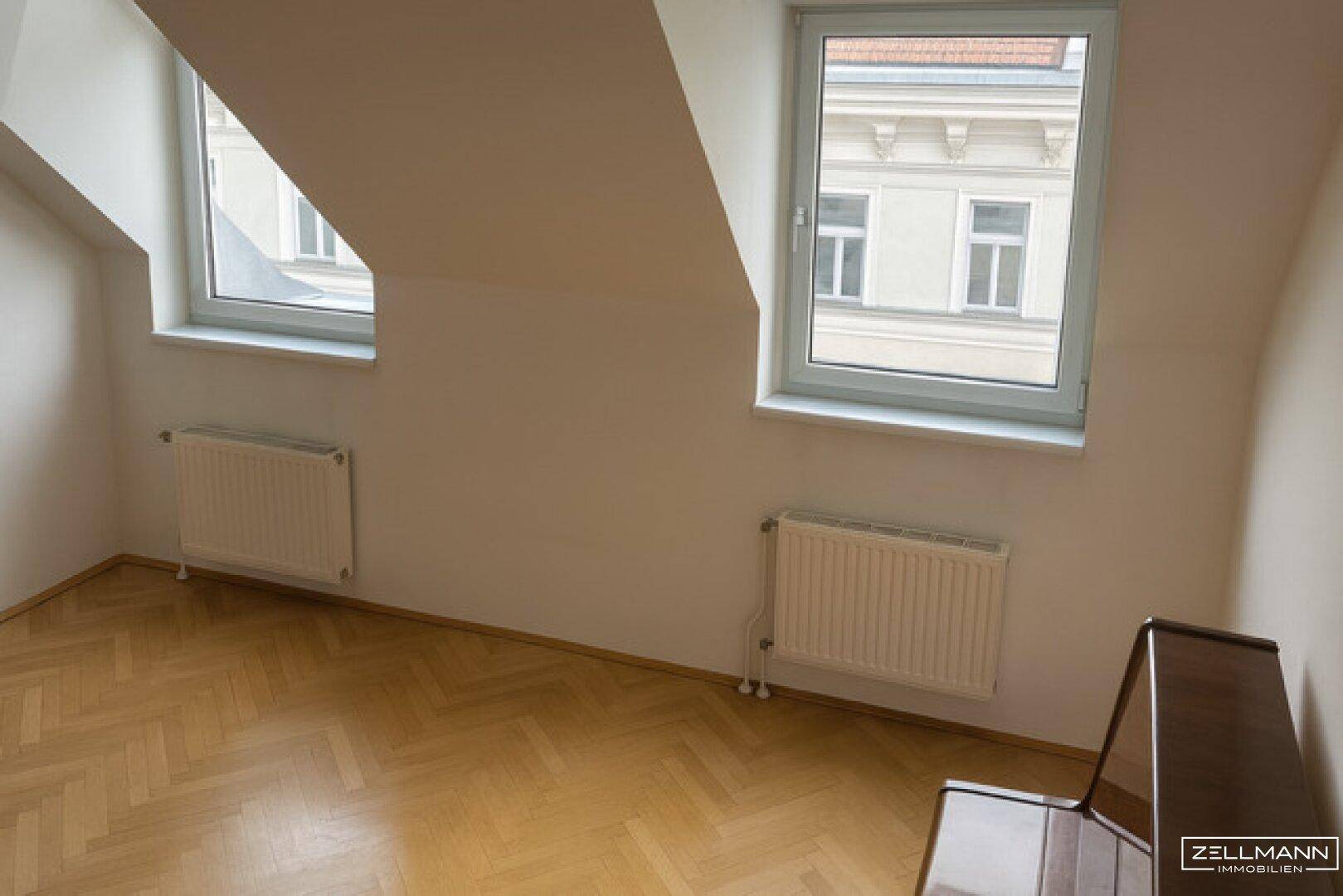 Dachgeschosszimmer mit schrägen Wänden, zwei Fenstern und Heizkörpern.