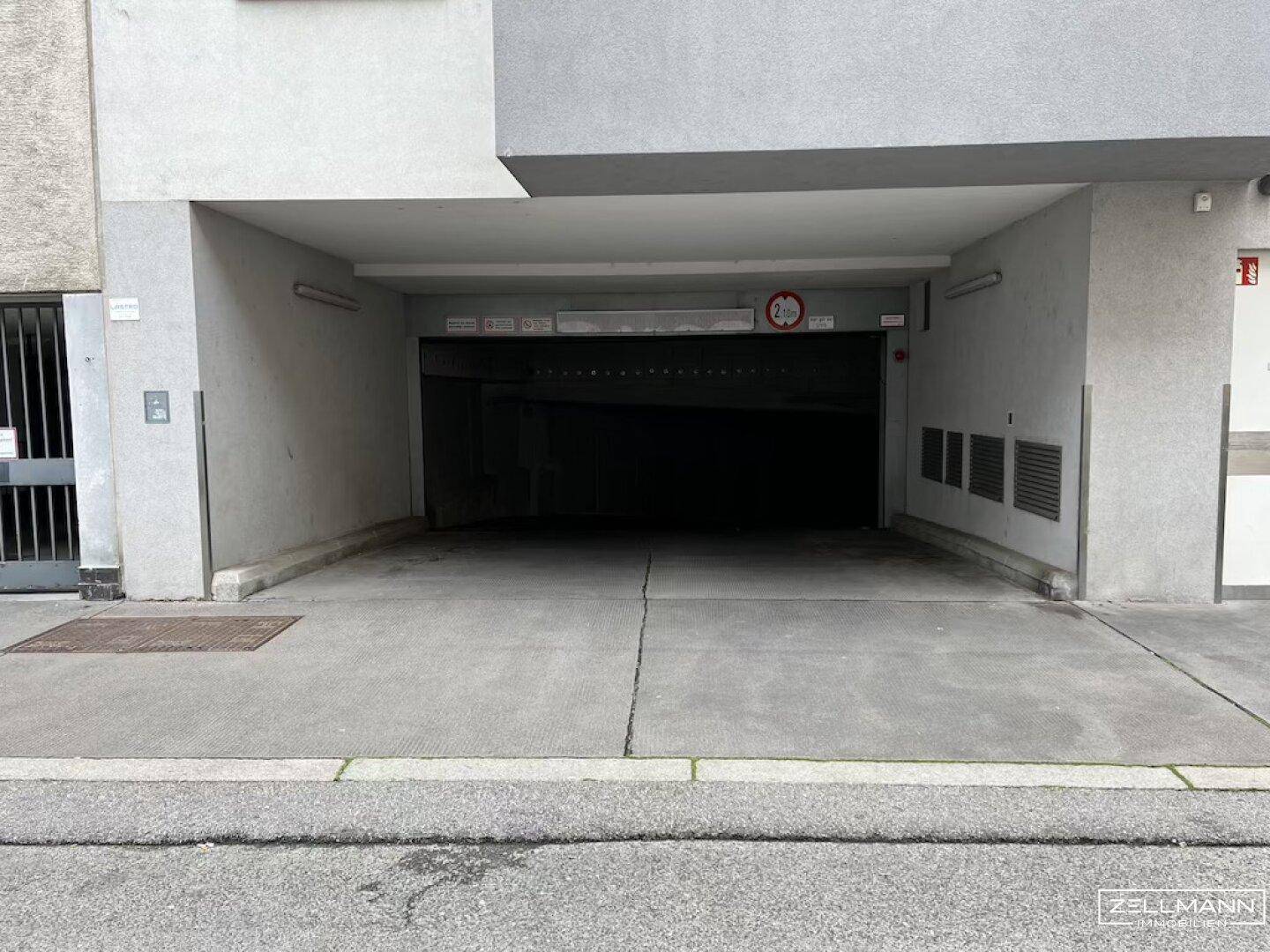 Einfahrt zu einer Tiefgarage mit Höhenbegrenzung und grauer Fassade des Gebäudes.