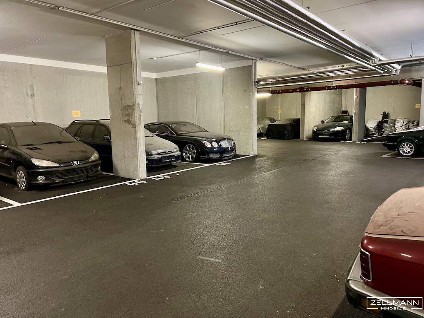 Weitläufige Tiefgarage mit zahlreichen geparkten Fahrzeugen und sichtbaren Versorgungsleitungen an der Decke.