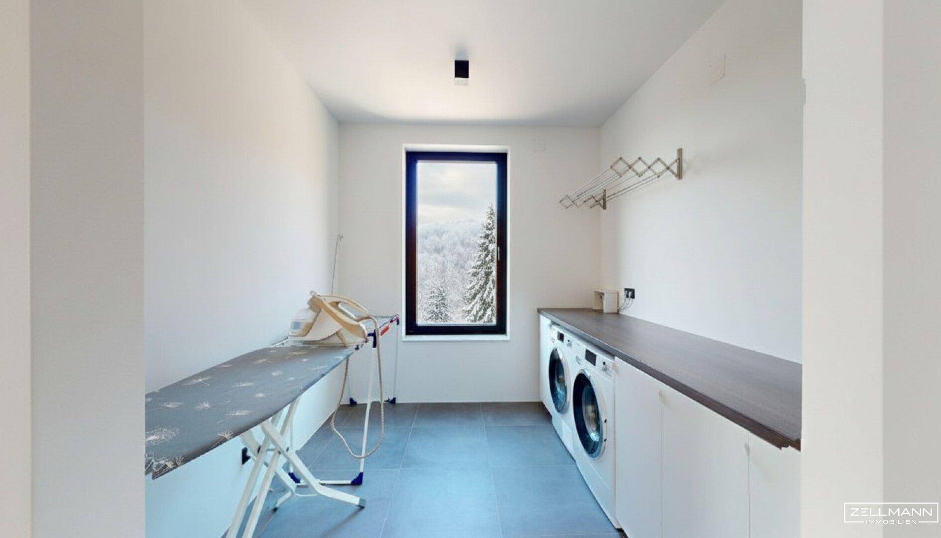 Funktionaler Hauswirtschaftsraum mit Waschmaschinen, Bügelbrett und einem Fenster mit Ausblick.