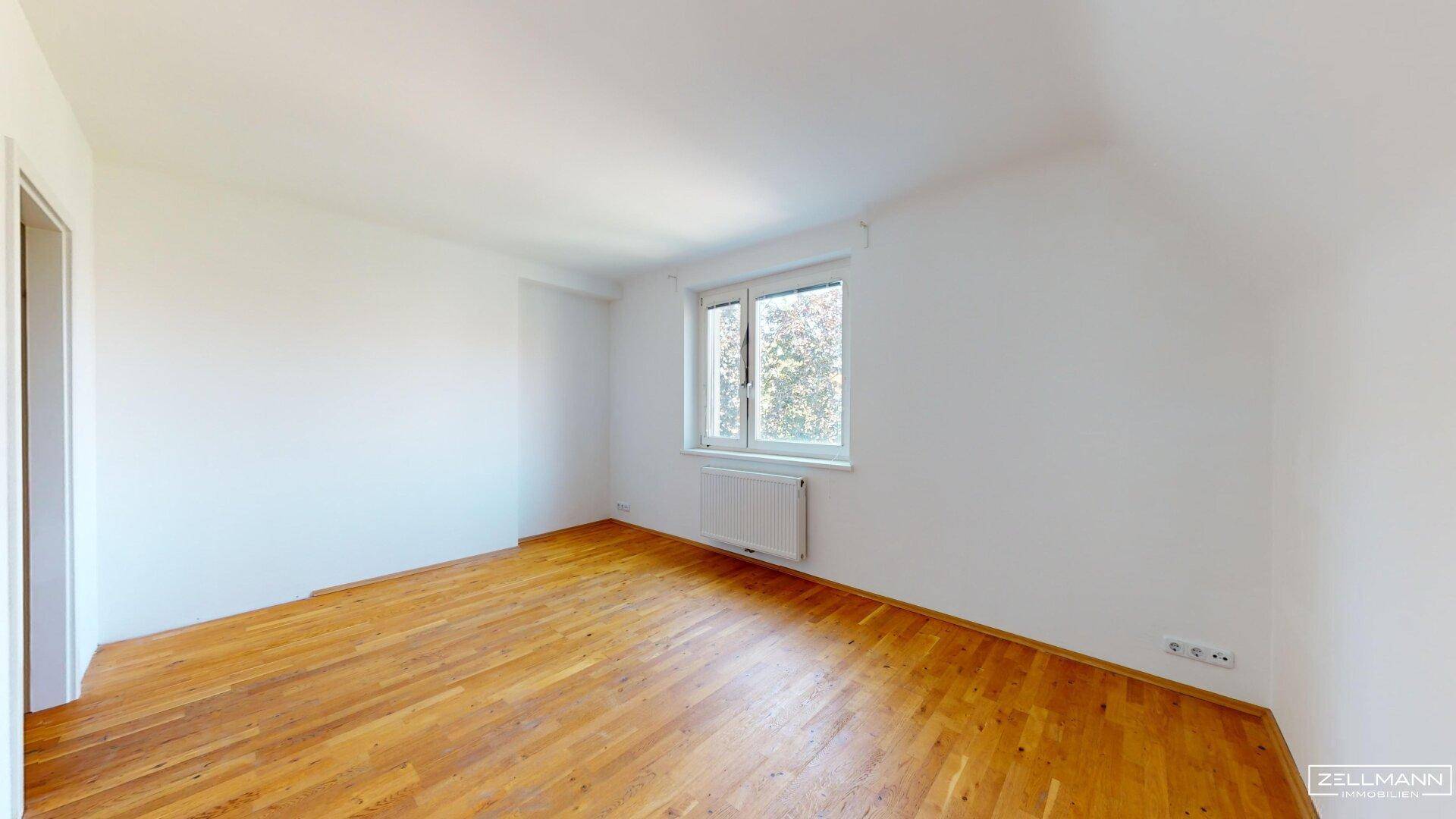 Helles, leeres Schlafzimmer mit Holzboden und einem Fenster, das den Raum mit Licht füllt.