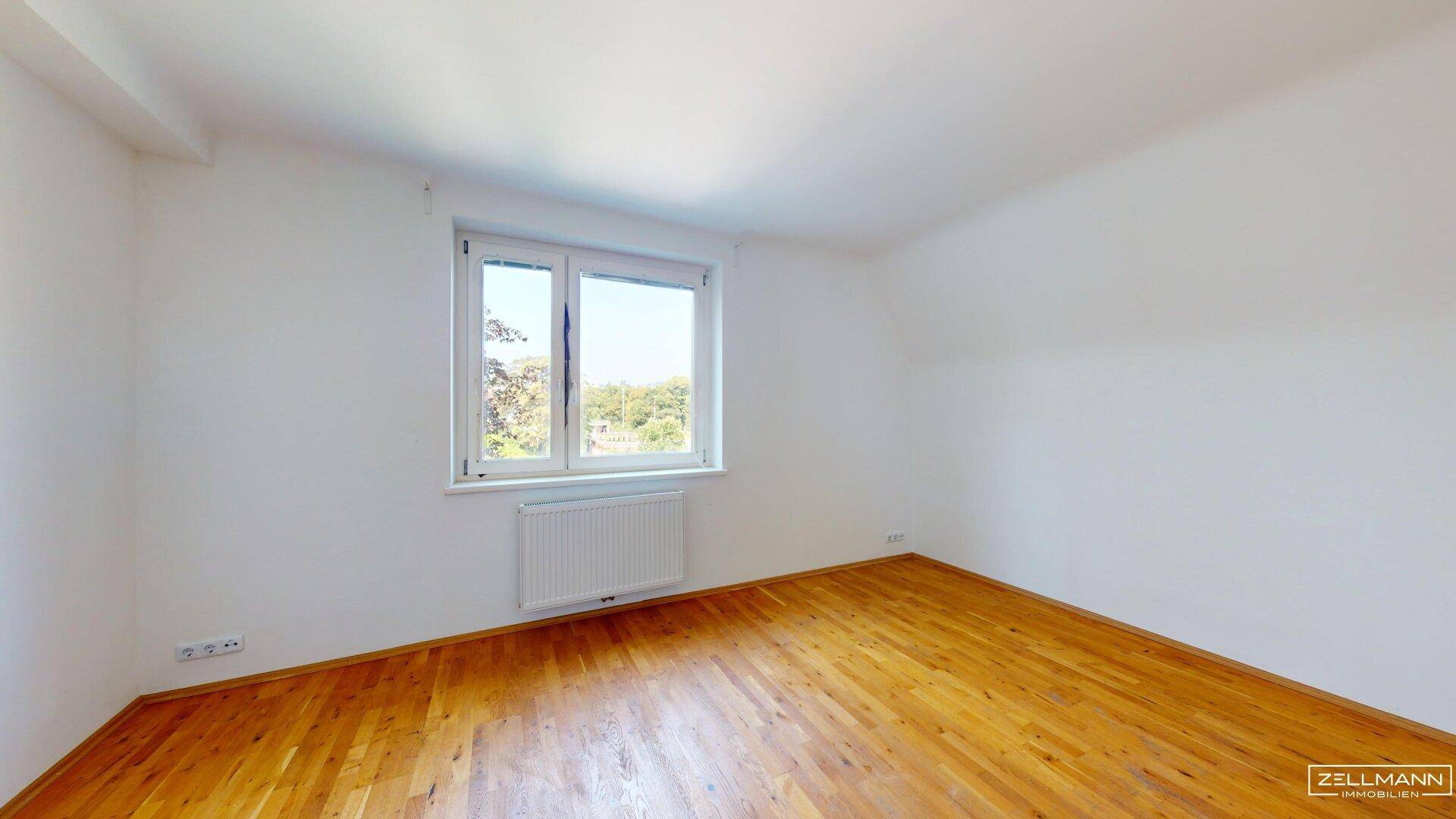 Geräumiges, leeres Schlafzimmer mit Holzboden und einem großen Fenster für viel Tageslicht.