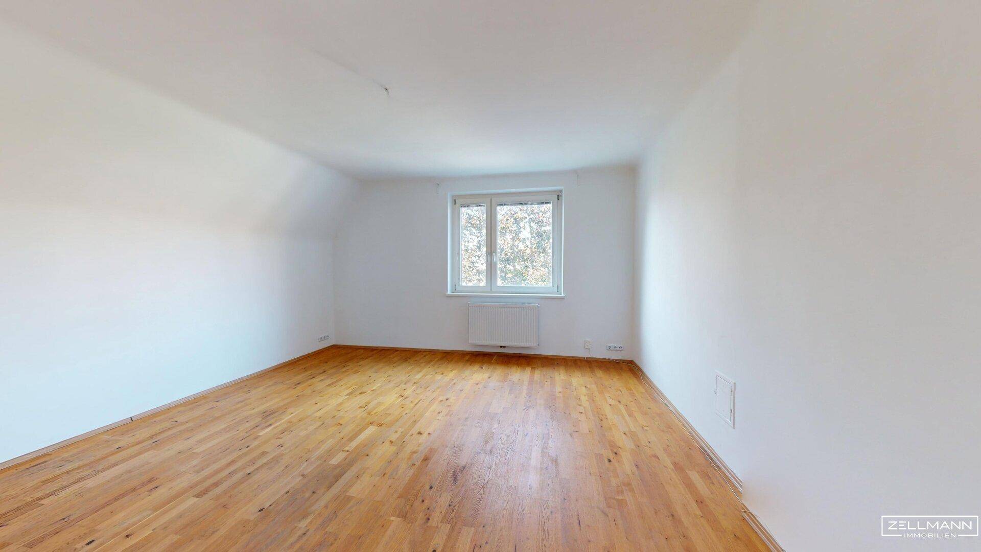 Helles, leeres Schlafzimmer mit Holzboden und einem Fenster, das viel Tageslicht hereinlässt.