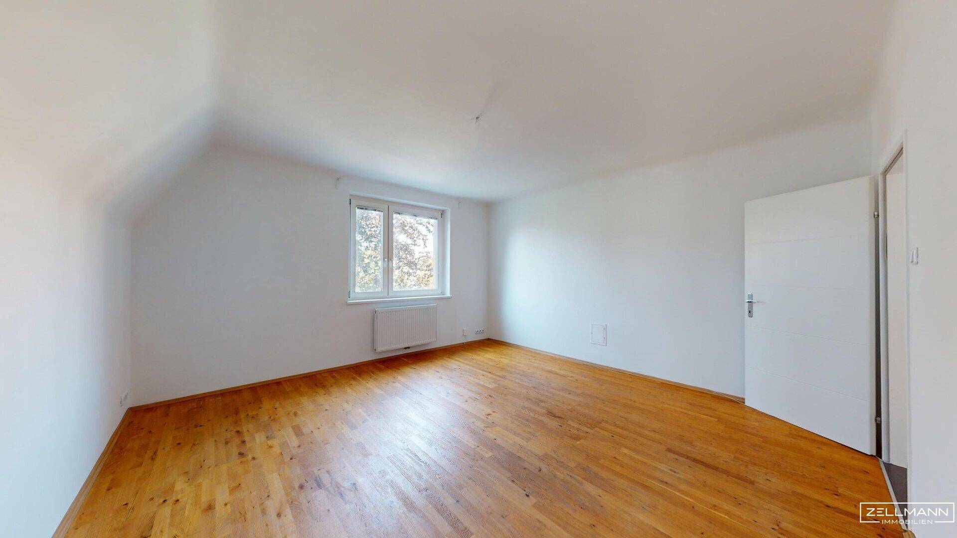 Leeres Schlafzimmer mit Holzboden, weißen Wänden und einem Fenster, das den Raum erhellt.