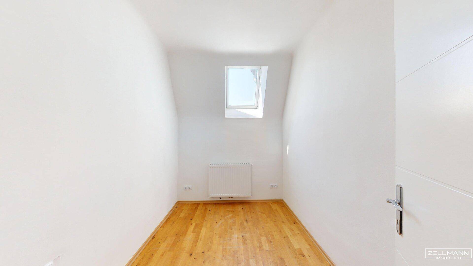 Kleines, helles Zimmer mit Holzboden, weißen Wänden und einem Dachfenster.