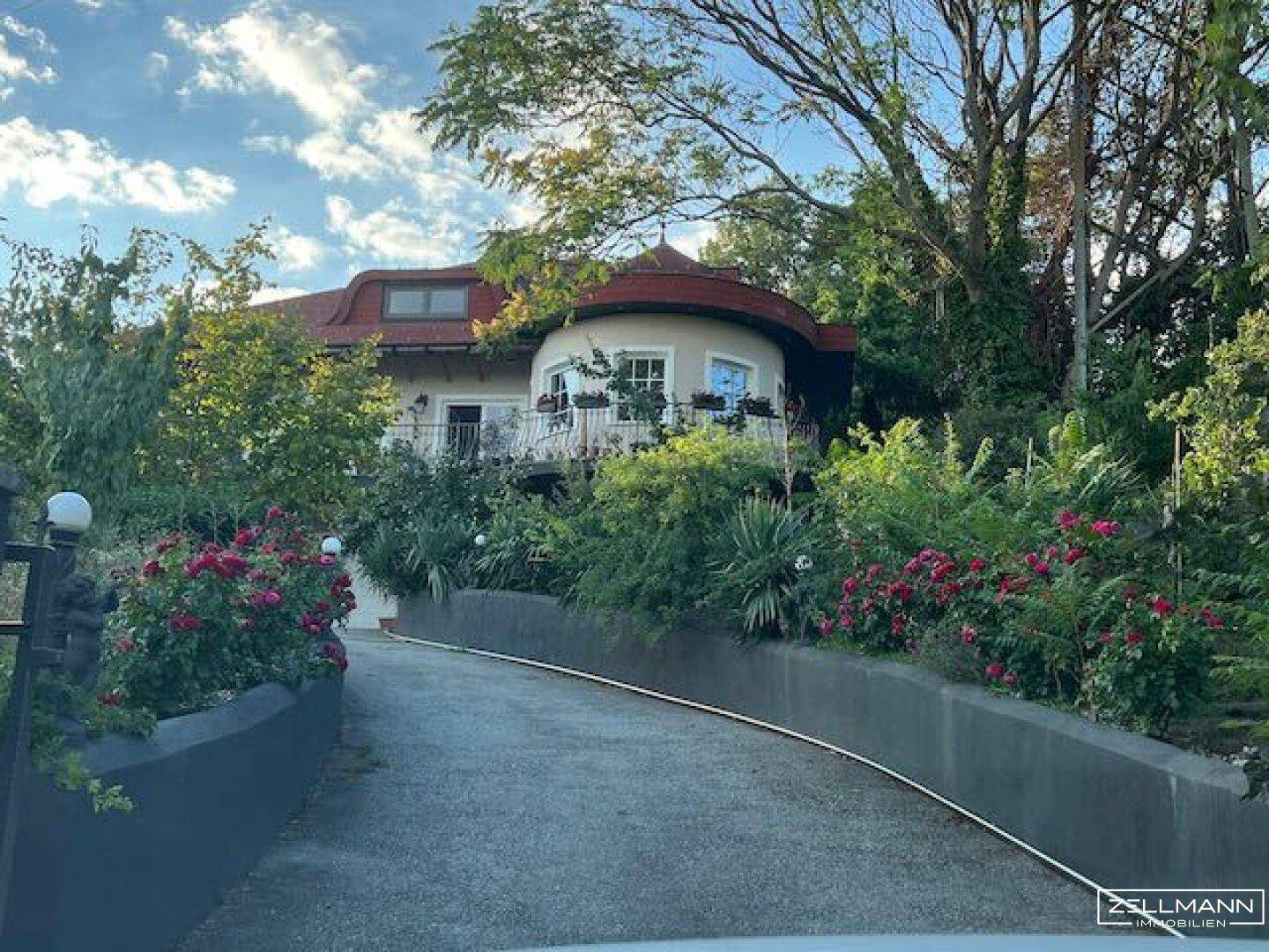 Elegante Villa mit geschwungenem Dach und Auffahrt, umgeben von einem blühenden Garten.