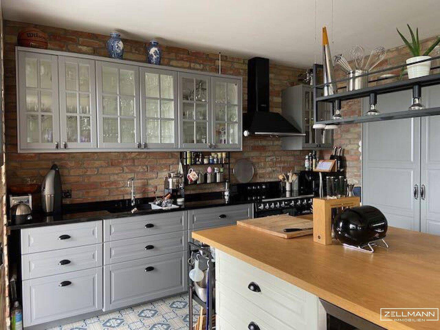 Detailansicht der Küche mit grauen Schränken, Backsteinwand und schwarzer Arbeitsplatte.