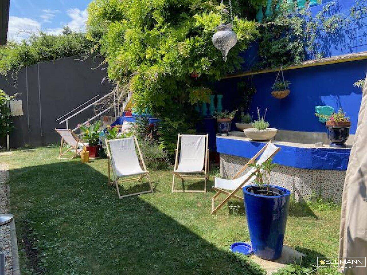 Mediterraner Gartenbereich mit Liegestühlen, blauer Wand und üppiger Bepflanzung.