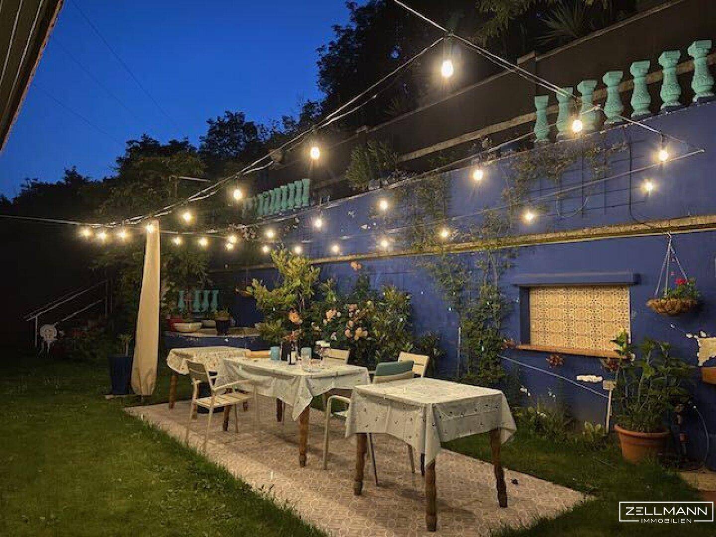 Abendliche Gartenterrasse mit Lichterkette, Esstischen und blauer Wand.