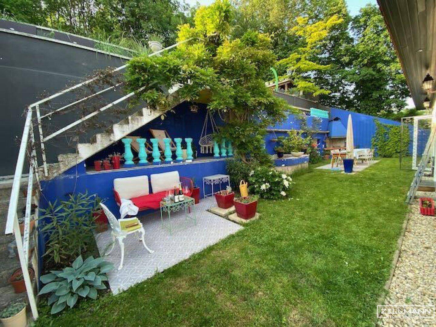 Mediterraner Gartenbereich mit Sitzgelegenheiten, blauer Wand und grüner Rasenfläche.