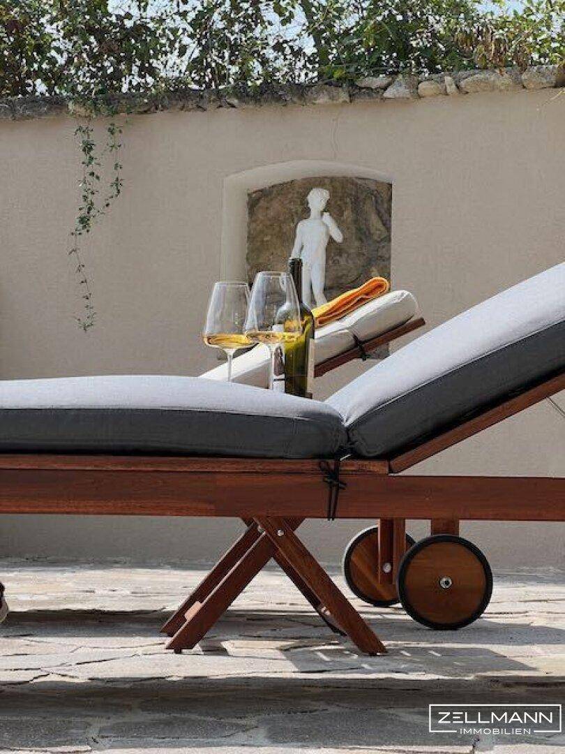 Sonnenliege am Poolbereich mit Blick auf eine Statue und mediterrane Pflanzen.