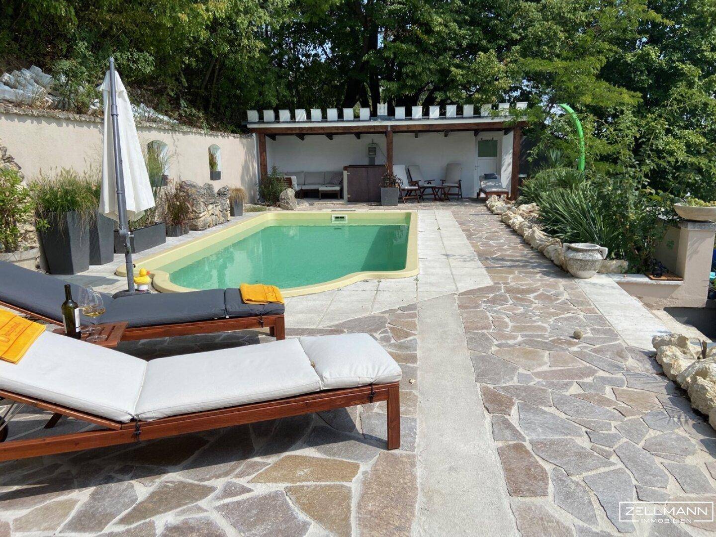 Großer Außenpoolbereich mit Sonnenliegen, Pergola und gepflasterter Terrasse.