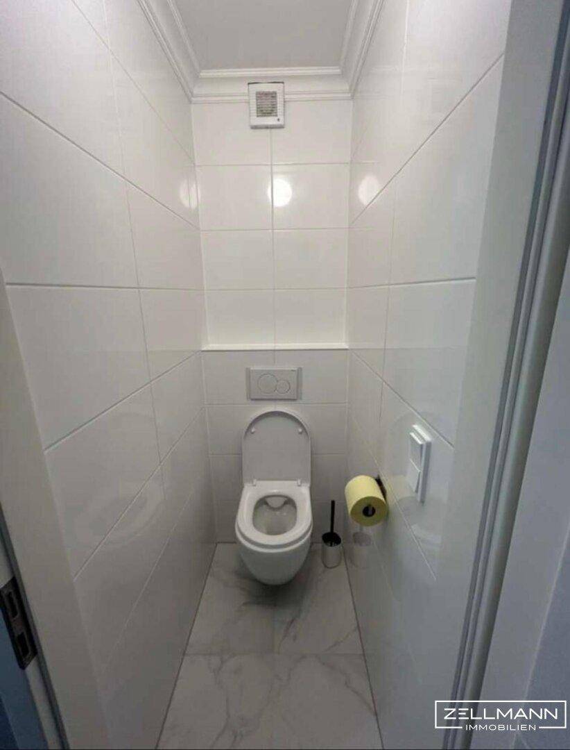 Kleines, hell gefliestes Gäste-WC mit Wand-WC und Lüftung.