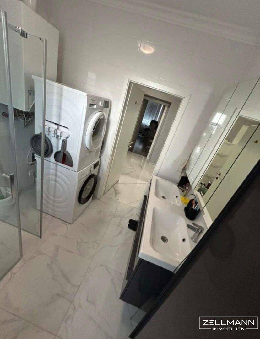 Helles Badezimmer mit Doppelwaschbecken, Spiegelschrank und integrierter Waschmaschine.