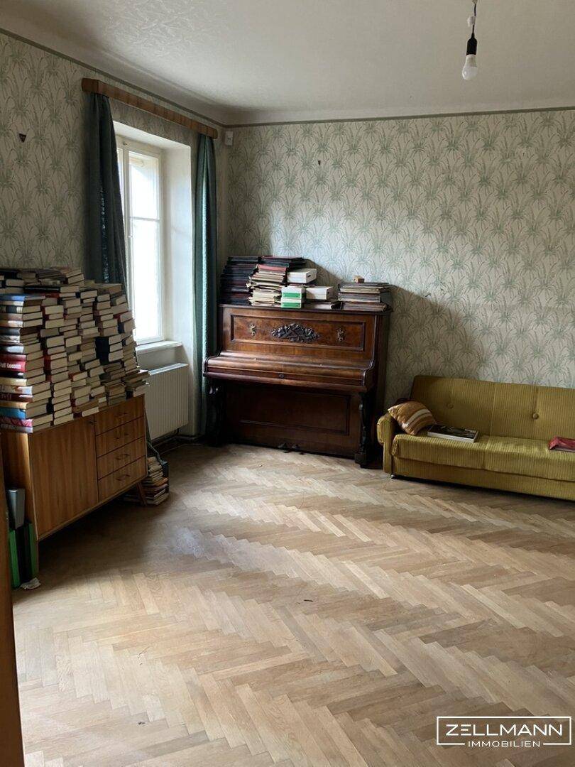 Helles Zimmer mit Fischgrätparkett, Klavier und einer gelben Couch, umgeben von Büchern.