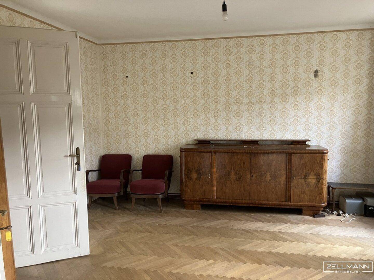 Geräumiges Zimmer mit Fischgrätparkett, gemusterter Tapete und einem antiken Sideboard.