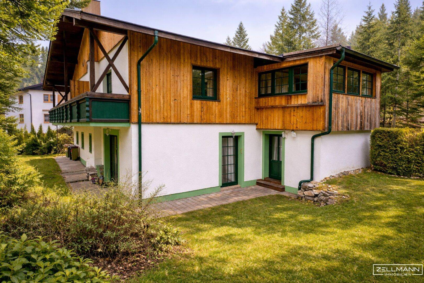 Rückansicht des Holzhauses mit Balkon, Terrasse und weitläufigem Garten, umgeben von Bäumen.