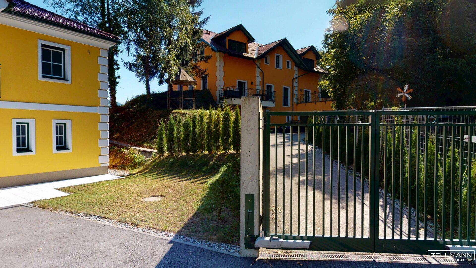 Einfahrt zum Anwesen mit grünem Tor und gepflegtem Vorgarten, umgeben von Bäumen.