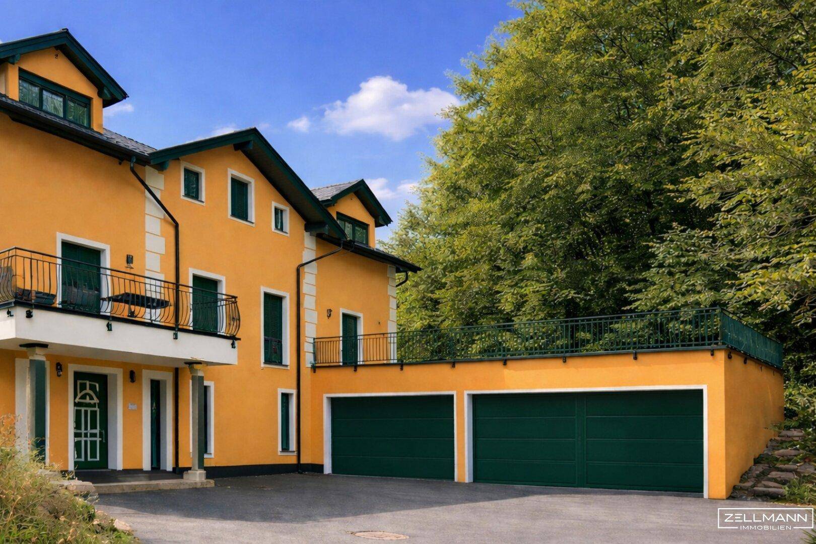 Gelbes Einfamilienhaus mit grünen Garagentoren und Balkonen, umgeben von viel Grün.