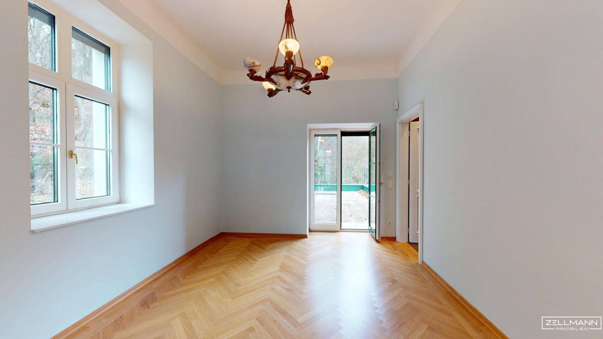 Helles Zimmer mit Fischgrätparkett, großen Fenstern und Zugang zu einem Balkon.