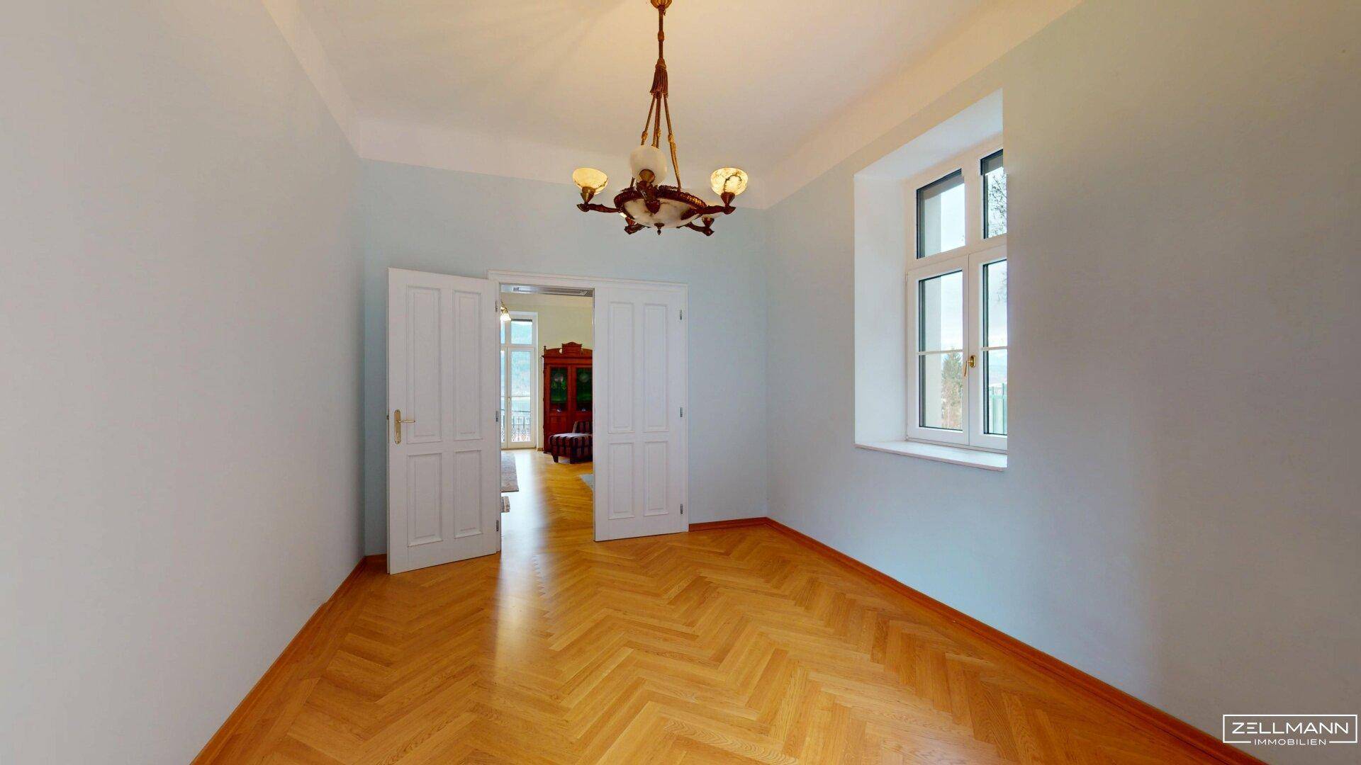 Geräumiges Zimmer mit Fischgrätparkett, großen Fenstern und Doppeltüren.