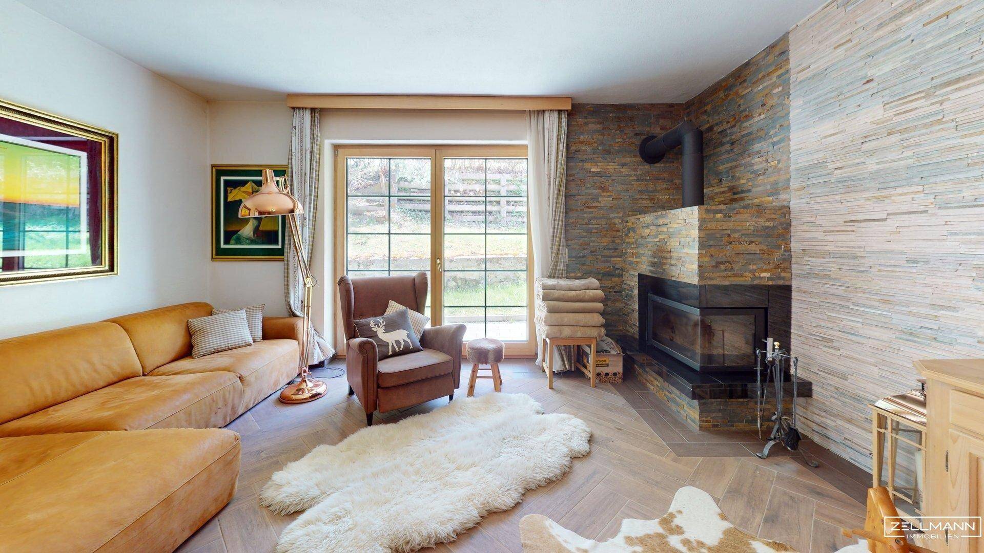 Gemütliches Wohnzimmer mit Kamin, Ledersofa und großen Fenstern, die viel Tageslicht hereinlassen.