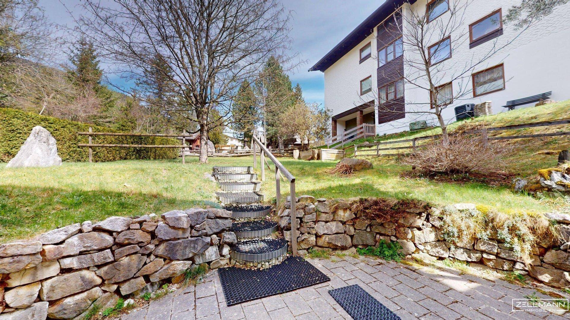 Gepflasterter Außenbereich mit Steintreppe und Rasenfläche, umgeben von Bäumen und Natur.